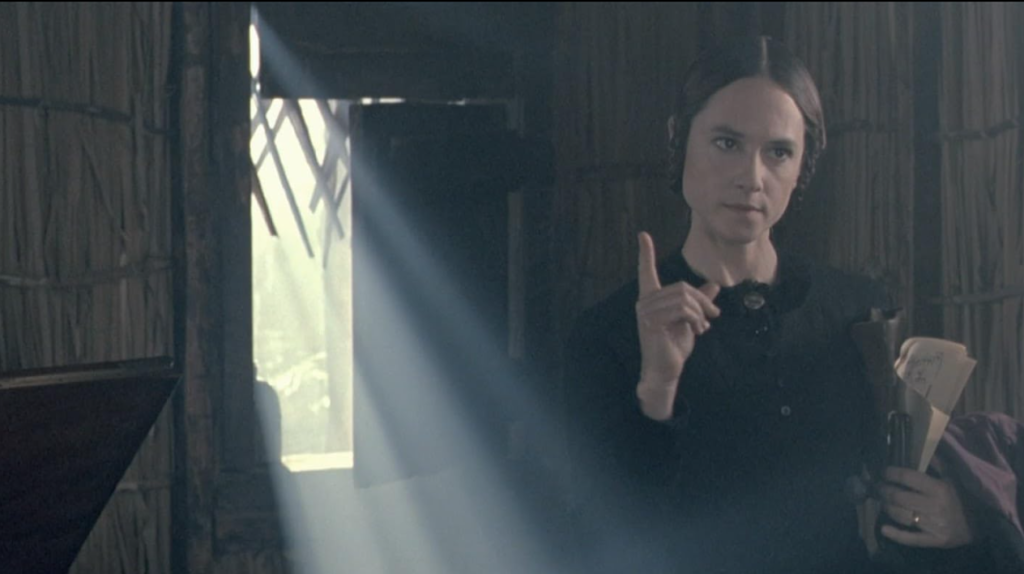 Ada stands in a darkened room with one finger extended toward the camera.