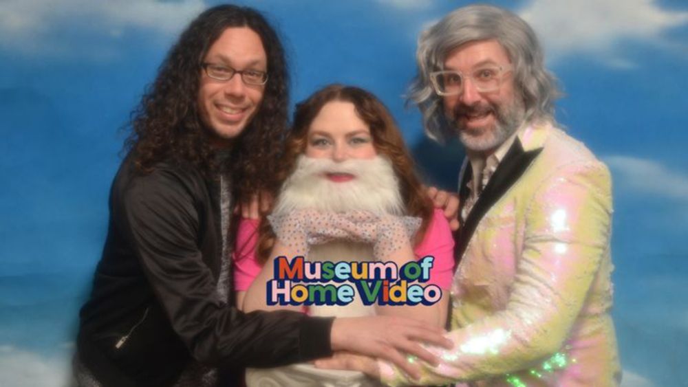 Three individuals standing in front of a sky-and-cloud-themed portrait background. The light-skinned man on the left has curly long brown hair and wards glasses, the light-skinned woman in the middle has red hair and is wearing a fake white bears, the light-skinned man on the right is wearing a grey wig and clear -framed glasses, and a sparkly formal suit.