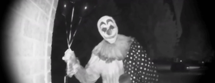 A blurry black and white image by a door camera, showing a person dressed as a scary clown, holding three balloons, standing in someone's doorway, facing the camera.