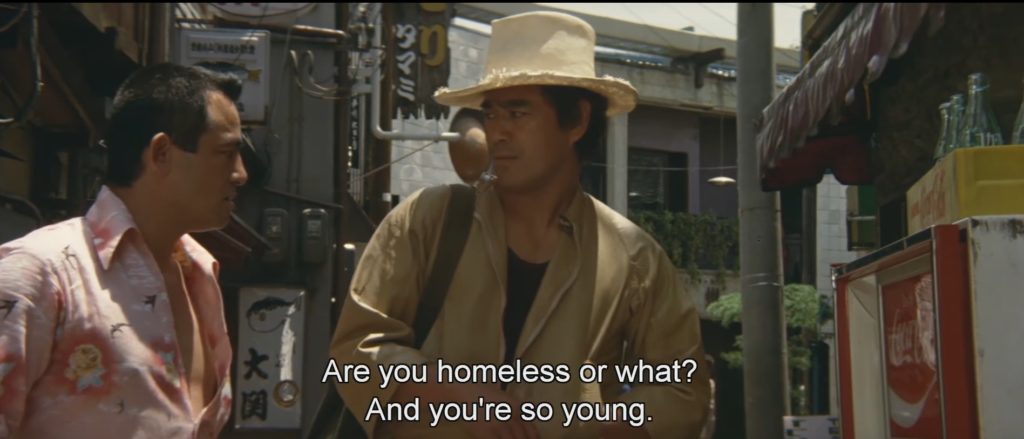 A shopkeeper in a pink shirt asks a skeptical Joji Kano, “Are you homeless or what? And you’re so young.”