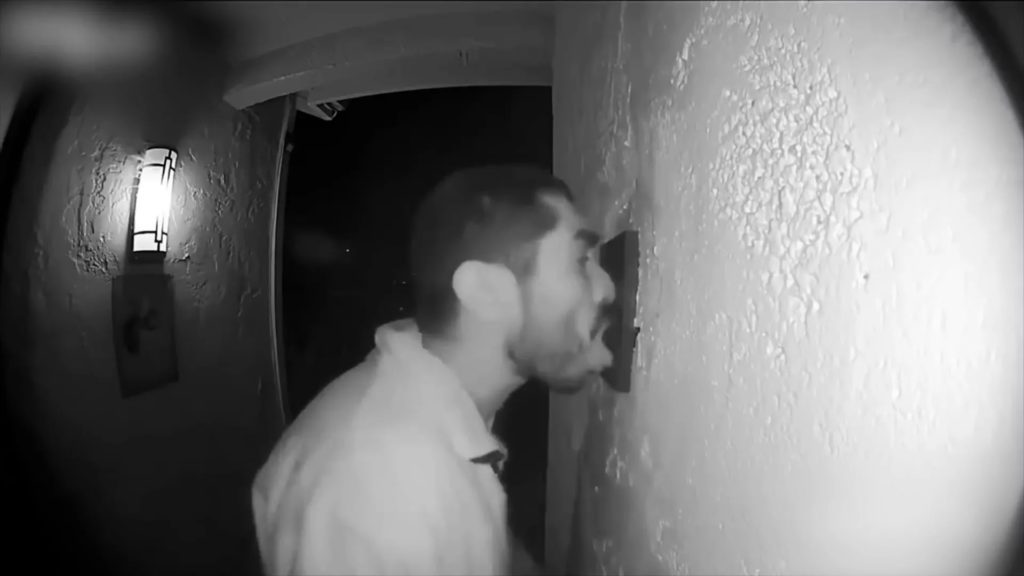 A blurry black and white image of a door camera, showing a man at night licking part of the entry way wall.