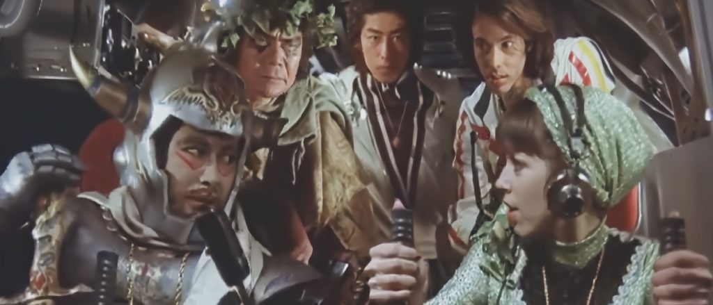 Five people gather in the cockpit of a spaceship. Chiba wears silver armor and red makeup around his eyes; he leans over to talk to the pilot.