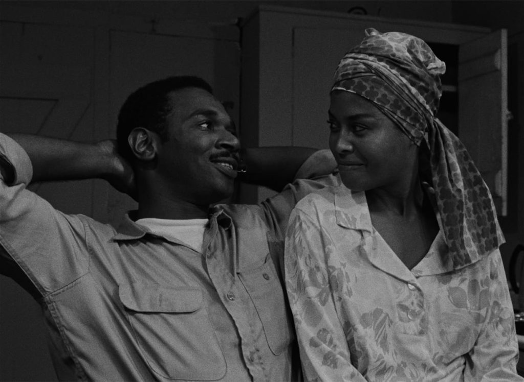 Ivan Dixon as Duff, a young Black man with short hair and a mustache, is sitting with his arms up behind his head, smiling at Abbey Lincoln playin Josie, a Black woman with a patterned scarf around her hair, who reciprocates his smiling glance.