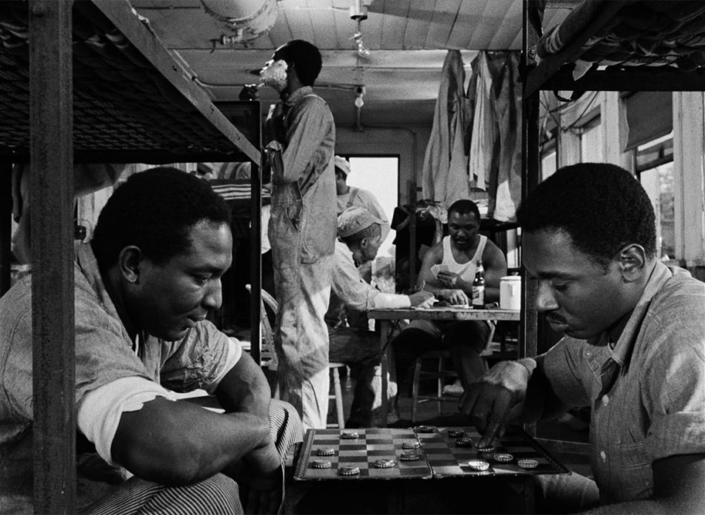 Duff is sitting on a bunk bed with a Black co-worker, playing checkers with beer caps. Many other Black men populate the background of the room, shaving, playing cards, or standing around.