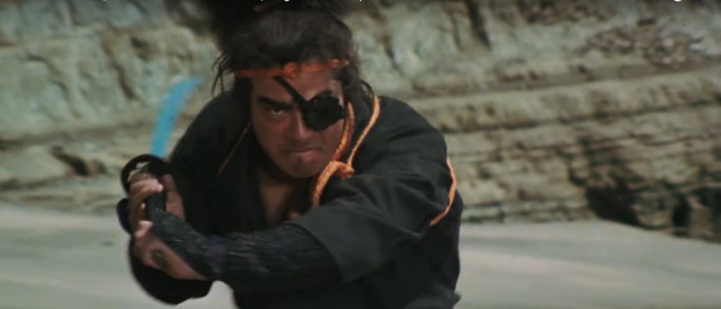 Chiba is seen running along a beach toward the viewer; he wears an eyepatch and brandishes a sword.
