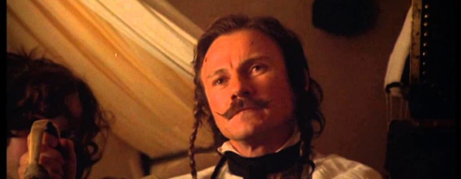 Feraud (Harvey Keitel), a light-skinned, brown-haired man with a mustache and two skinny hair braids, is sitting down with a person on his right whose face is obscured by Feraud's hand. He is wearing a white shirt with a thick black collar, and looking slightly off-camera.