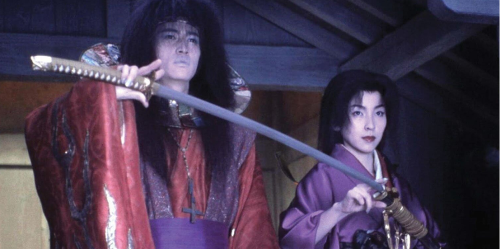A man in an ornate red and purple robe holds a gold hilted sword with his right hand. He is standing to the right of a woman in purple kimono.