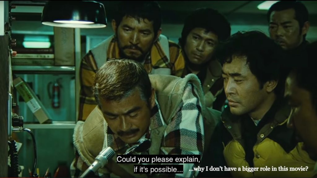  Chiba and other actors gather around a CB radio. The subtitle reads “Could you please explain, if possible…” Added to the image are the words “...why I don’t have a bigger role in this movie?”