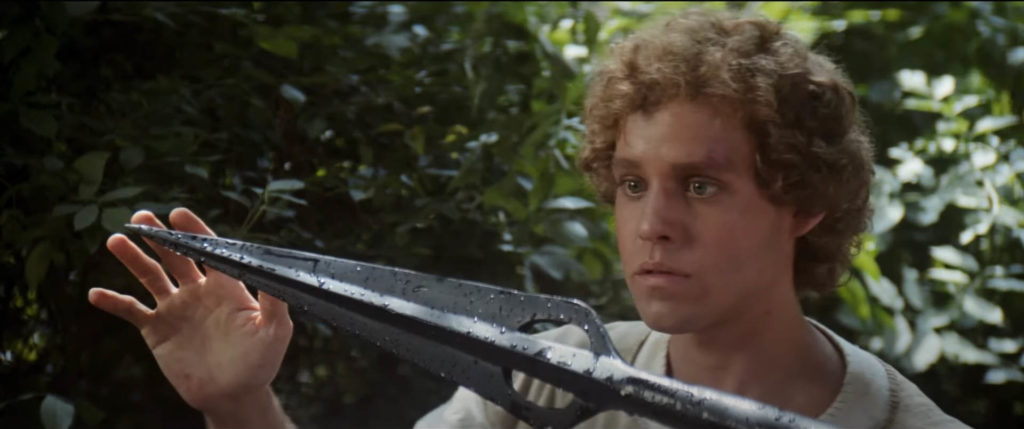 Peter MacNicol as the protagonist Galen Branwardyn looks in awe at the tip of a massive metal spear, reaching out to touch it with his right hand.