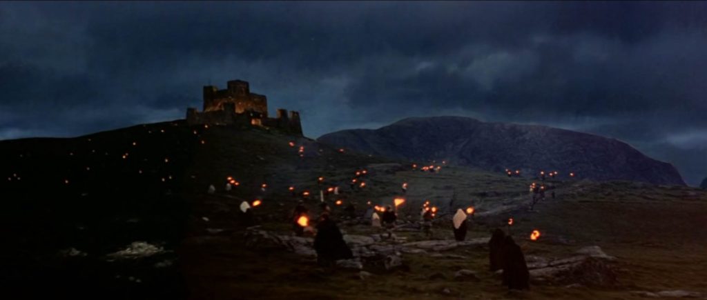 A moody stone castle on top of a hill underneath a dark overcast sky. In the foreground dozens of small figures walk towards the castle holding torches.