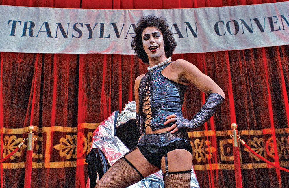 a white man with black curly hair wearing a pearl necklace, a black corset, black underwear, black gloves, and stockings
