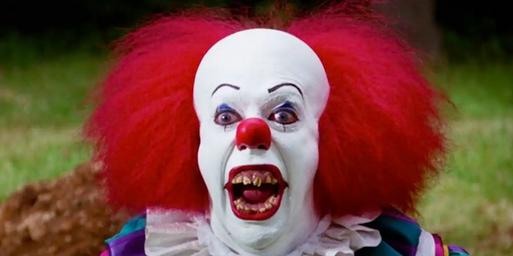 a man with bright red hair wearing white clown makeup with a red nose and with jagged teeth
