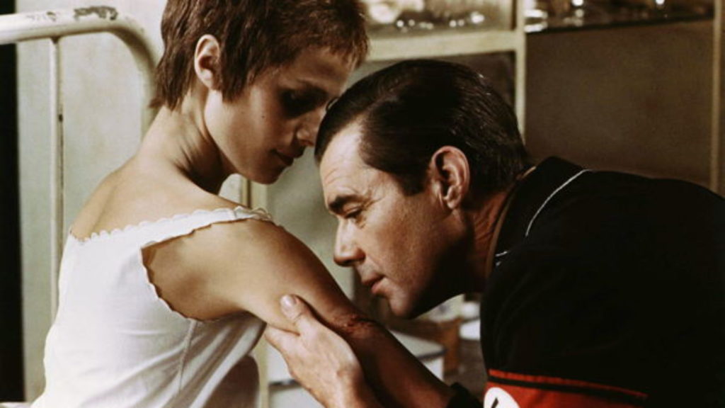 Charlotte Rampling as Lucia, a young woman with light skin and very short brown hair, is sitting on a white metal frame bed. Dirk Borgade, as Max, an older light-skinned man with dark, sleek hair, and a nazi uniform, is leaning into Lucia’s wounded arm, appearing to be on the brink of kissing it.