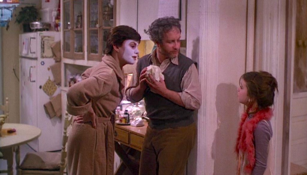 Marsha Mason as Paula McFadden, a light-skinned women with short brown hair, wearing a bath robe and a cosmetic face mask, stands next to, Richard Dreyfuss as Elliot Garfield, a light-skinned man with short brown hair and a salt & peppered beard, leaning against the kitchen door frame. They both face Quinn Cummings as Lucy McFadden, Paula’s daughter, wearing a fuzzy red vest.