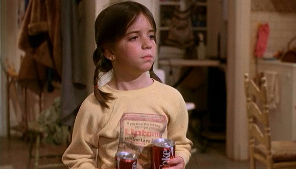 Quinn Cummings as Lucy McFadden, Paula’s daughter with medium brown braided hair, is holding two cans of coke in her hands, wearing a light-yellow sweater.