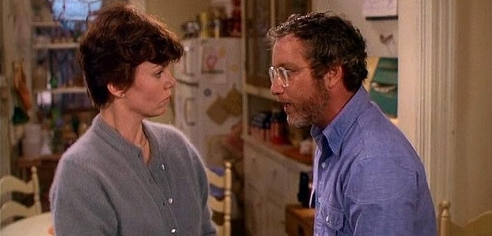 Marsha Mason as Paula McFadden, a light-skinned women with short brown hair, wearing a grey wool cardigan, stands on the left side of a kitchen, facing Richard Dreyfuss as Elliot Garfield, a light-skinned man with short brown hair, glasses, and a salt & peppered beard, who is reciprocating her look. 