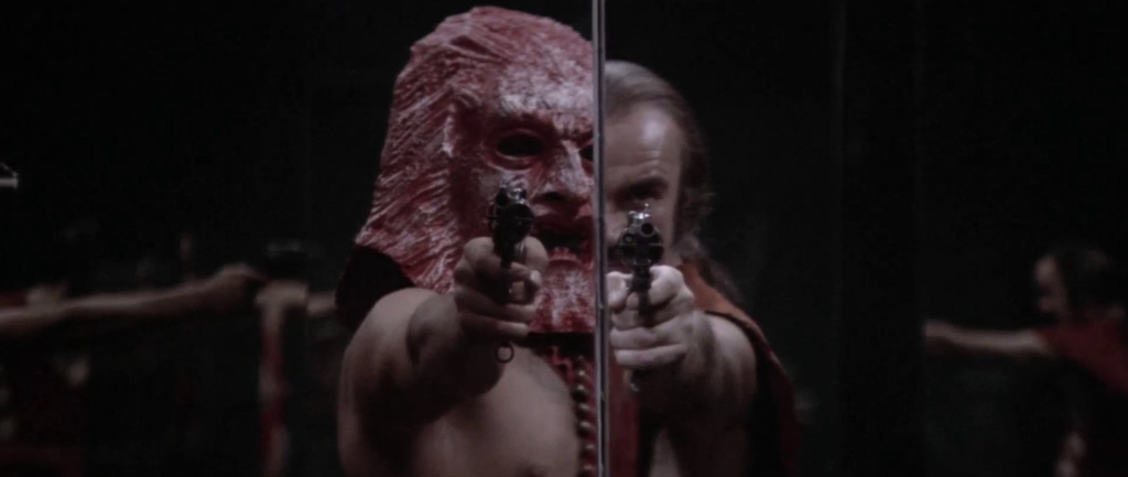 A man with long hair and a mustache fires a handgun in a hall of mirrors as a man wearing a large mask does the same