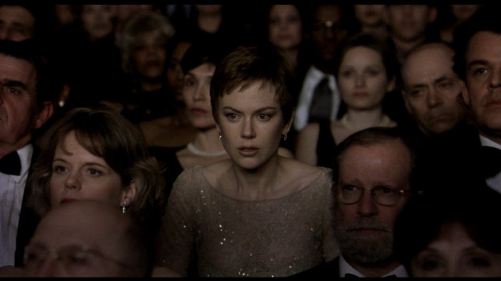 A closeup of Nicole Kidman sitting in a dimly lit theater, surrounded by well dressed people