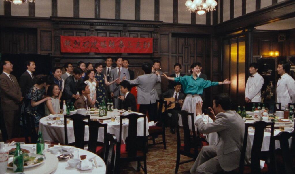 Multiple people are spread out across the frame at a banquet, all in dress attire, in a dining hall with dark wood paneling. Food and drinks are spread out across the white tablecloths, and all the guests are in the middle of song. Two figures toward the center of the frame, a man in a gray suit and a woman in a teal blazer, have their arms extended, guiding the melody. On the wall is a red banner with kanji characters.