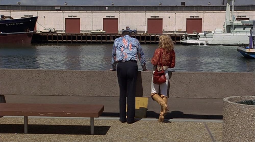George C. Scott and Season Hubley stand in a wideshot, their backs to us, overlooking a dock, with a building facing them and a few boats parked on the water in between. Scott has black pants and a long sleeved blue collared shirt with a pink design on it, and has his hands resting on the railing with a posture that’s slightly hunched over. Hubley has blonde shoulder length wavy hair, a dark orange jacket and purse, tight white pants and tan knee-high boots. A bench and a trash can are in the foreground.