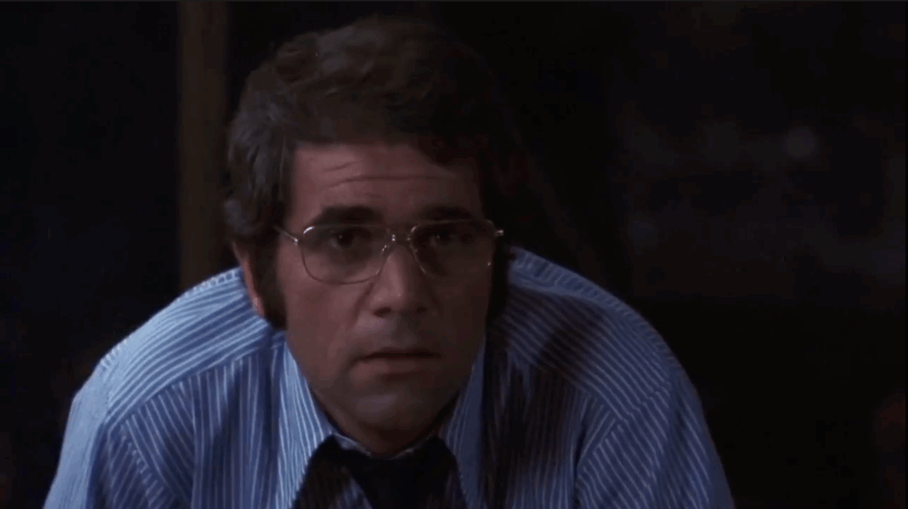 A man in glasses looks intently ahead. The man is hunched over slightly with a concerned look on his face.