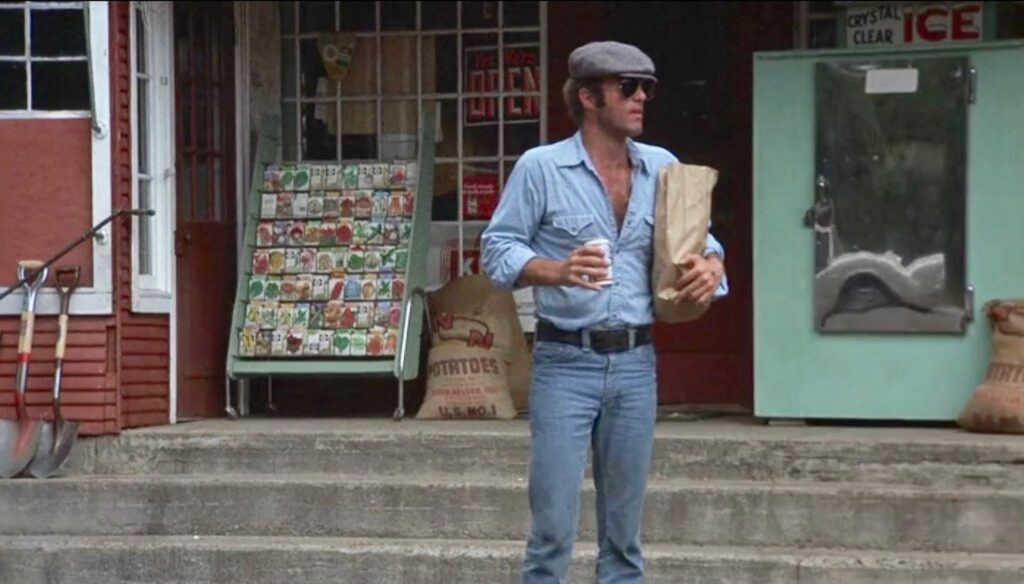 A man stands in front of a store holding a cup of coffee and a brown paper bag. He is wearing a denim blue shirt, a hat, and large sunglasses.