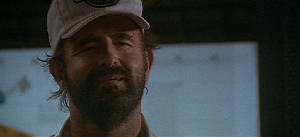 An off-center headshot of a grimacing man wearing a white baseball cap. His hair and beard are unkempt.