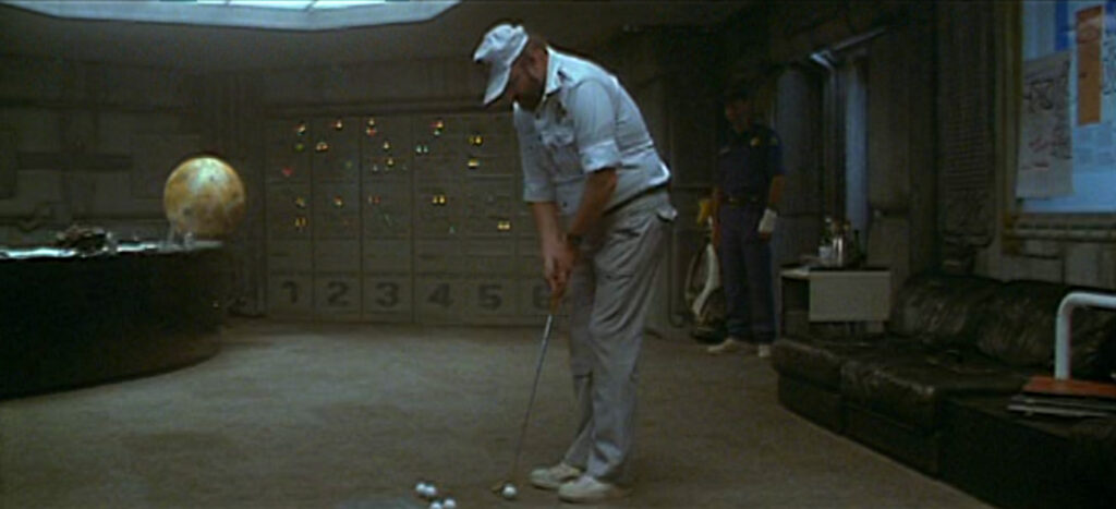 A man dressed in golf whites practices putting golf balls in an office. The office features a leather sofa, a model of Jupiter on a desk, and a flashing light board. In the background, a mand ressed in blue watches.