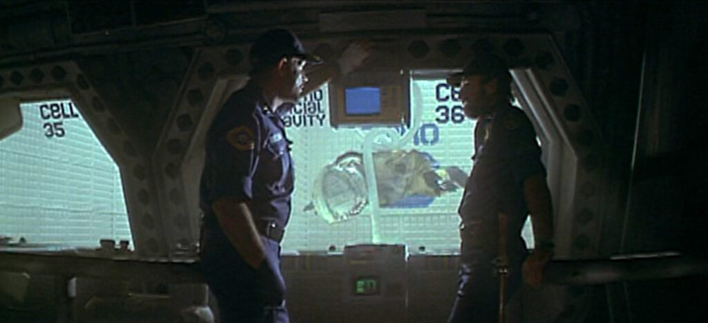 Two men dressed in blue talk. Behind them, a man wearing a space suit floats face down in a security cell.