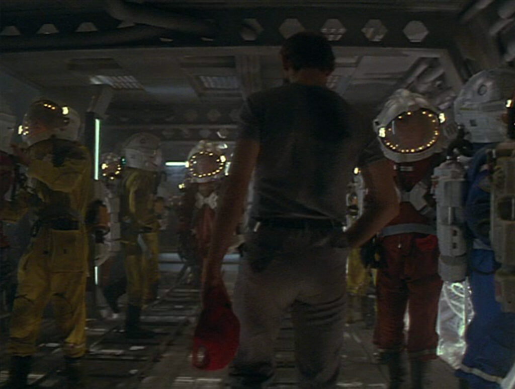 A man dressed in a t-shirt and pants carries a red baseball cap while he walks down a hallway populated by people who are all wearing spacesuits.