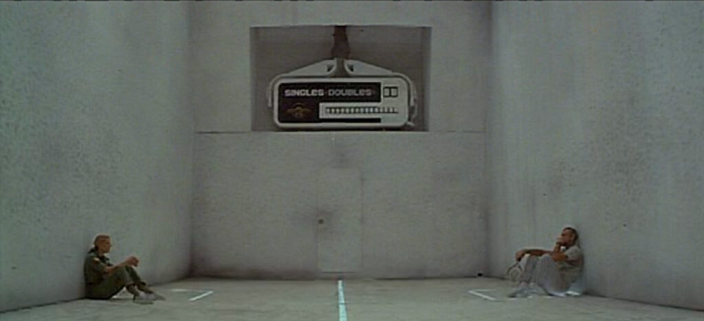 A wide shot of a racquetball court. On the left, a woman sits facing a man who sits on the other side of the court facing her.