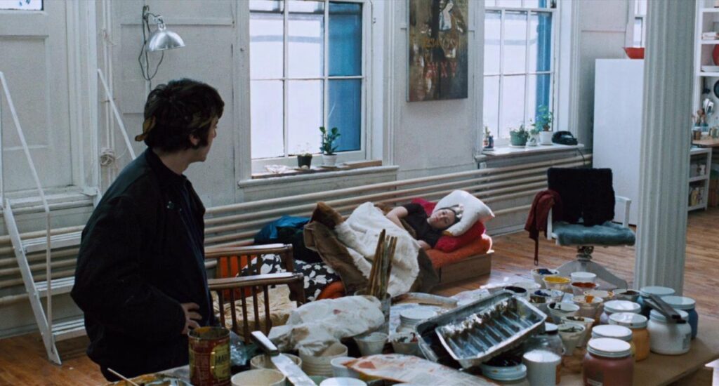 A man standing and a woman lying on the floor in a cluttered apartment.]