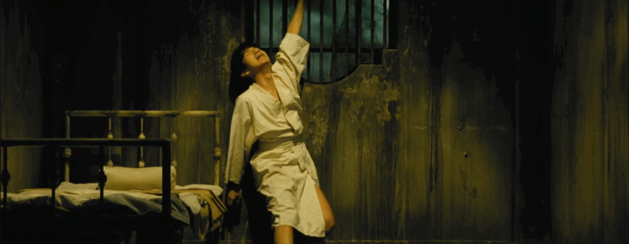 A light-skinned Asian man with short dark hair in a white robe is holding on to the bars of an ornamental window places on the wall of a dark room with faded walls and a minimally equipped bed placed on the left side of the frame. His face signals devastation.
