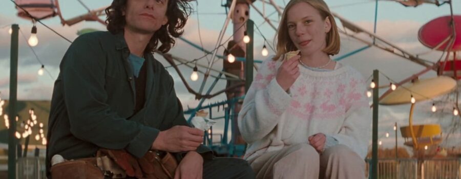 A medium-long shot of Tom McCamus as Sam Burnell sitting on top of a picnic bench alongside Sarah Polley as his daughter Nicole Burnell. Sam has one leg propped up on the bench of the table, his hands resting in his lap and holding a half-eaten ice cream. He's dressed in forest green coveralls and wearing a brown leather toolbelt, staring up and to the left with an inscrutable expression on his face, his shoulder-length dark hair blowing in the wind. Nicole has both legs propped up on the same bench and is dressed in beige, white, and pink clothing, including a patterned sweater with rabbits strewn across the chest. She is mid-bite of her cone and looks amused. The brightly lit background indicates they are at a carnival at dusk.