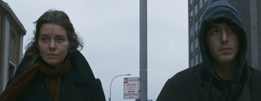 A woman and man are walking outside, the woman on the left and the man with his hood up on the right. there is a large apartment building behind them.
