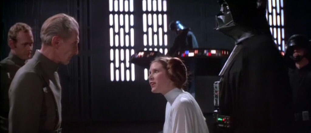 A small, young woman, Princess Leia, harangues two military men while a creature in monstrous black armor passively watches. In the background, soldiers work at a brightly lit control console.