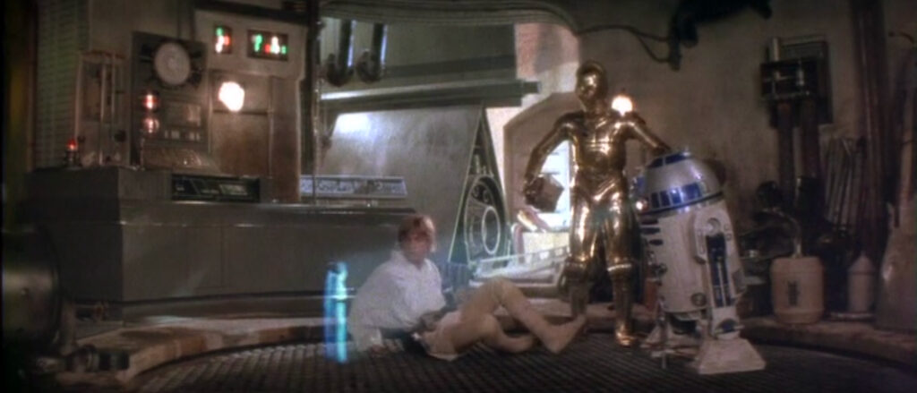 A surprised man, Luke, sprawls on the ground stares at a translucent image of a young woman, Leia. The image is projected by a squat blue-and-white droid while a tall golden droid watches.