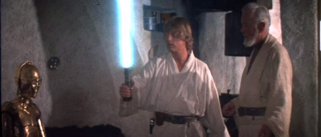 A young man, Luke, stares in awe at the lightsaber in his hand while an old man, Obi-Wan, watches. To their left sits a deactivated droid.