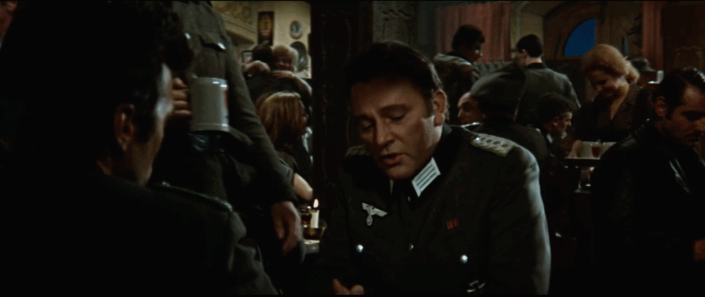 Two men sit facing each other in a crowded bar. One is looking at the camera while it sits over the shoulder of the other man)