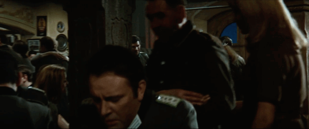 The camera has zoomed in slightly from the previous image. The man facing the camera is still in the frame but the camera is now focused on the door behind him. Other bar patrons can be seen in the background