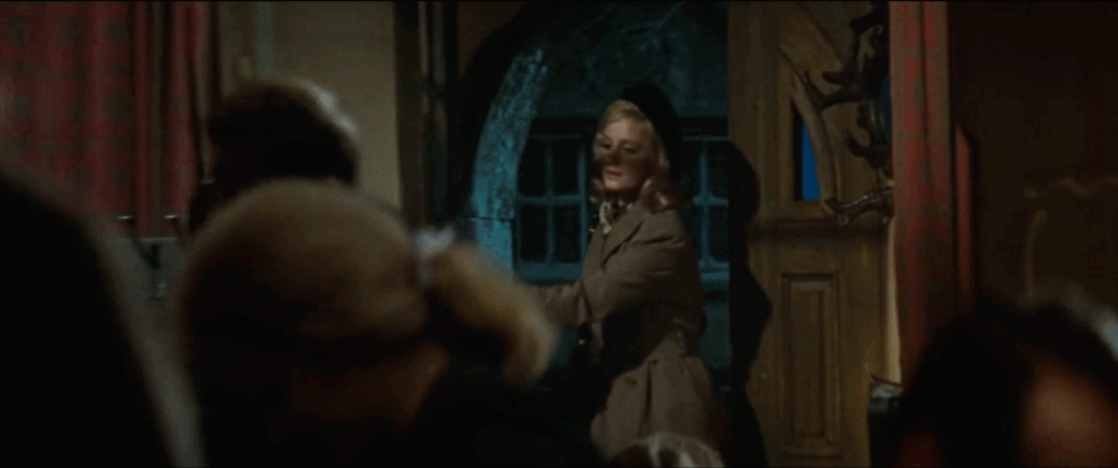 A woman opens the door and enters the bar. She is wearing a brown overcoat and black hat. She is carrying a purse in one hand)