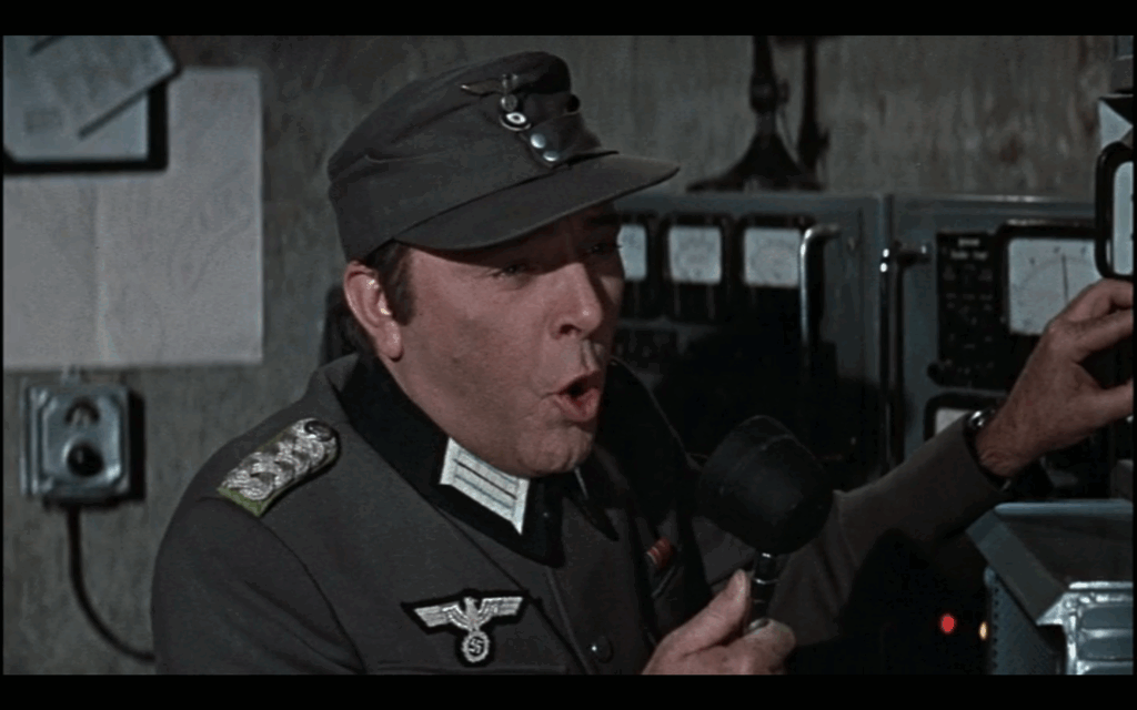 A man in a gray Nazi military uniform and hat speaks into a radio microphone, his mouth open in mid-speech.