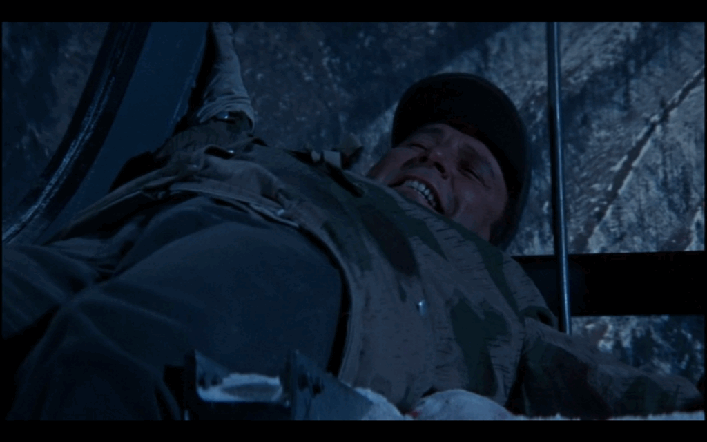 A man wearing a hat and straining his face lies on his back on top of a cable car.