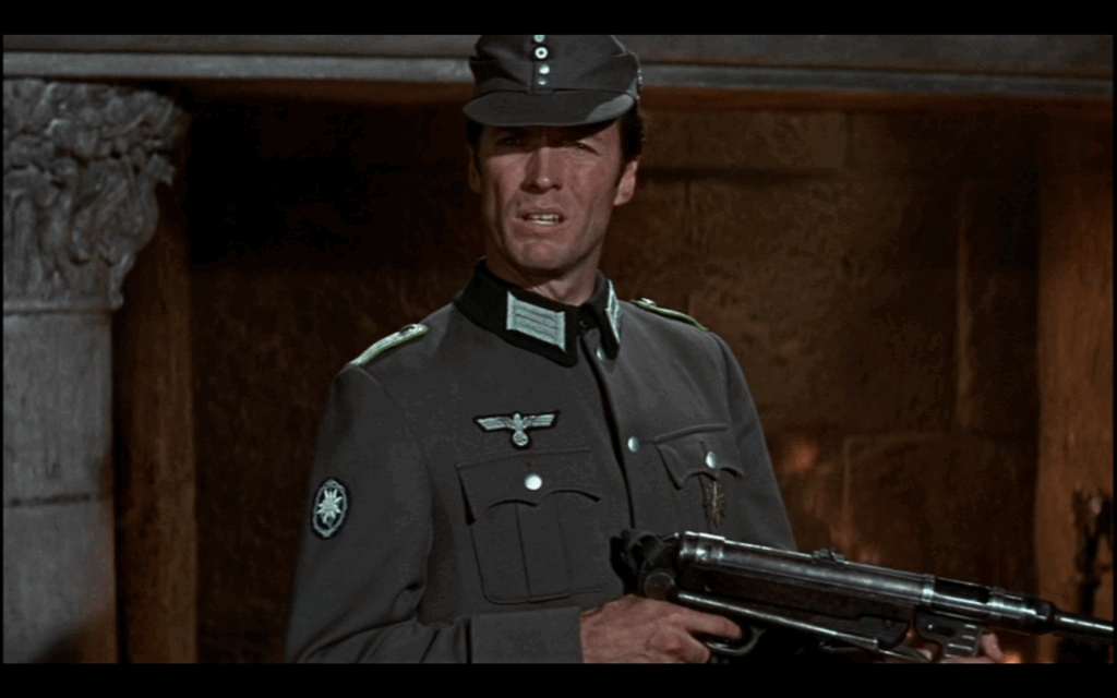 A man in a gray Nazi military uniform and hat holds a machine gun. He looks confused.