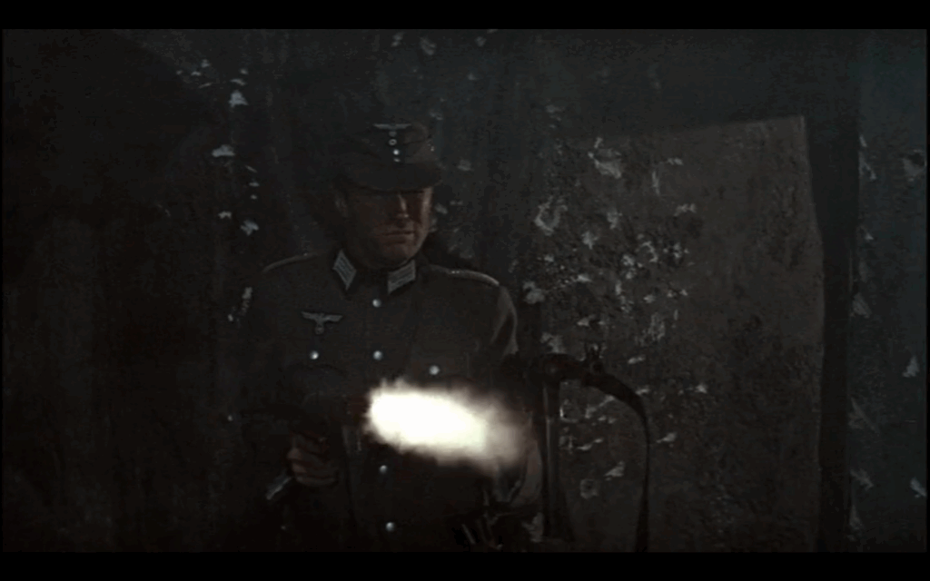 A man in a gray Nazi military uniform and hat shoots two big machine guns. I muzzle flash extends out from one of the guns.