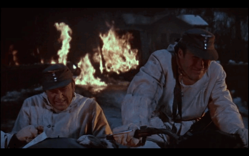Two men ride on a motorcycle. One drives and the other sits in the sidecar. A fire blazes behind them.