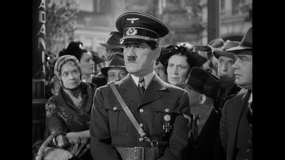 Black and white screenshot of a man in the foreground, shown from the waist up, who looks like Hitler in a military jacket and hat with white collar and tie, leather strap over one shoulder and belt. He is surrounded from behind by a crowd of people. One woman in particular leans in over his right shoulder, staring at him and wearing some kind of head wrap and holding a basket. Three men behind him and to his left are wearing fedoras, with a middle aged woman directly peering over his left shoulder. Everyone in the crowd is wearing something on their heads. Some out of focus buildings appear behind them.