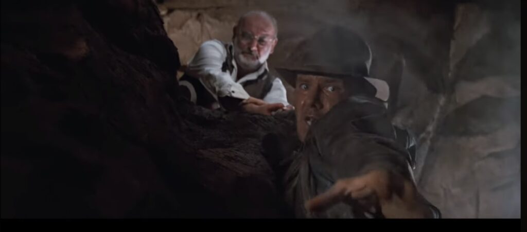Harrison Ford as Indiana Jones hangs from a precipice reaching for the Holy Grail, Sean Connery as Henry Jones holds onto Indy to keep him from falling.