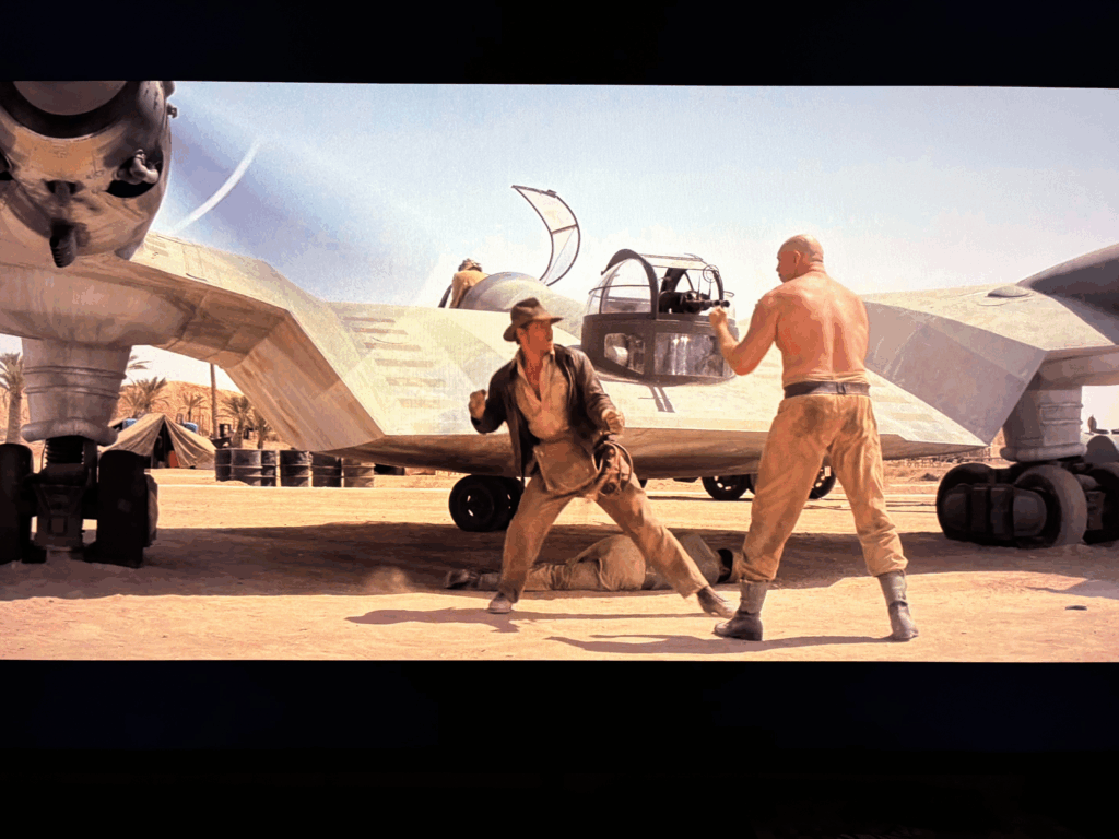 Harrison Ford as Indiana Jones draws back a mighty punch at a hulking shirtless Nazi in front of an airplane on a desert airfield.
