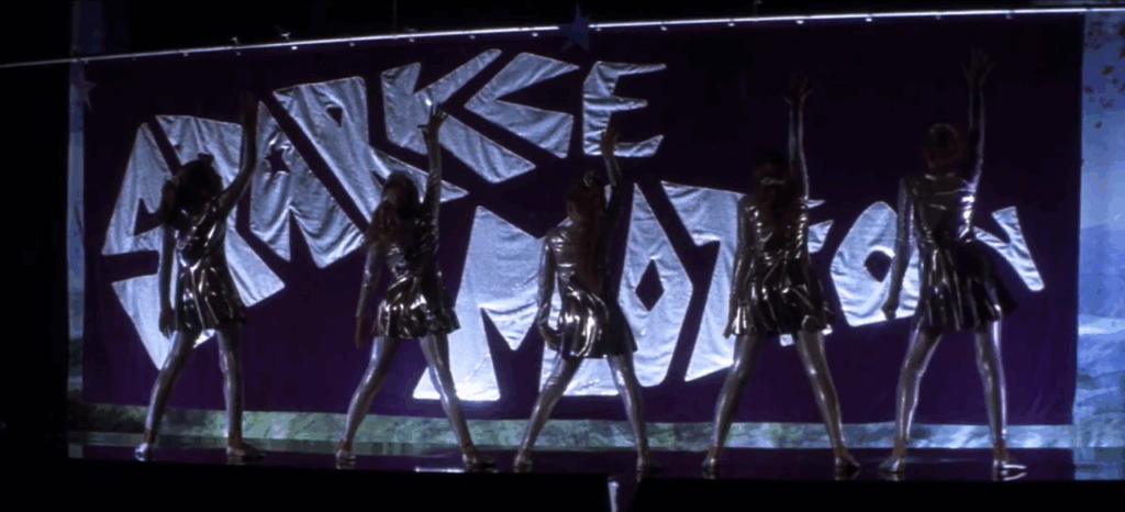 Five ten-year old dancers in reflective silver dresses, including Samantha Darko (Daveigh Chase) dance on stage, right arms raised, facing away from the audience towards a purple and silver sign which reads “Sparkle Motion.”
